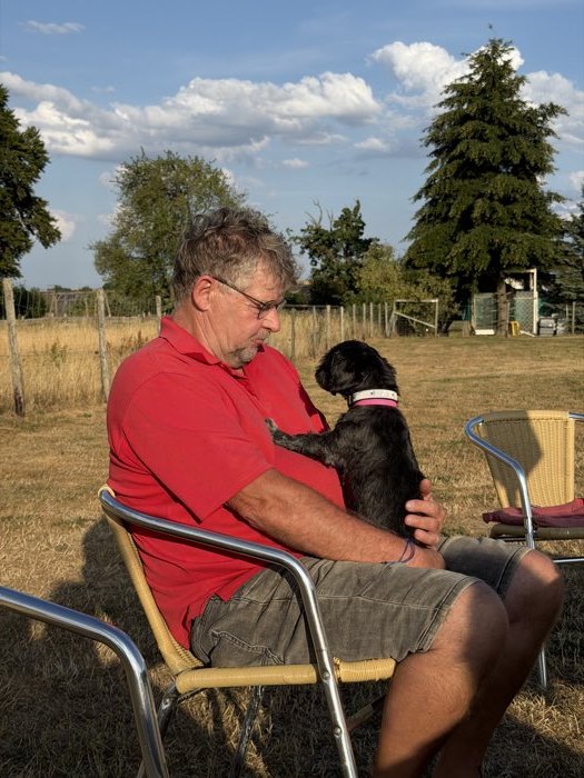 Martin Schmitz sitzt entspannt auf dem Hundeplatz, ein kleiner schwarzer Hund lehnt sich an ihn.